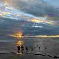 People walking out of the sea as the sun rises through the clouds behind them